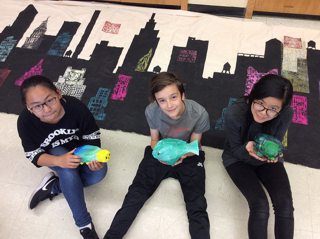 Students sitting on floor on top of city mural with papier mache fish