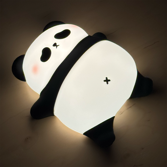A panda-shaped wall light glowing softly in a bedroom setting. The lamp features a cute cartoon panda design with black and white coloring, a small pink cheek accent, and a warm white illuminated body, mounted on a dark wood wall.
