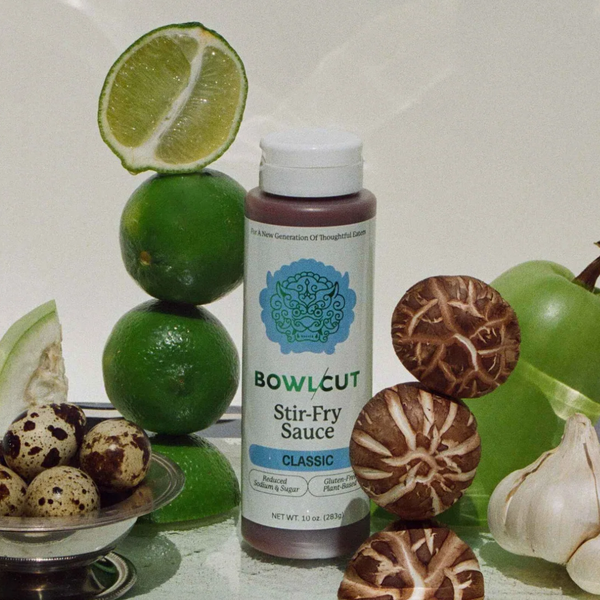 Bottle of Bowlcut Stir-Fry Sauce surrounded by limes, mushrooms, and garlic on a reflective surface.