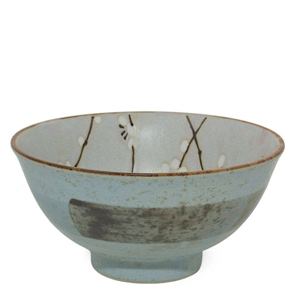 A handmade ceramic bowl with a light blue-gray glaze, featuring a rustic brown rim, a dark gray textured patch on the front, and delicate black branch and white blossom motifs painted inside.