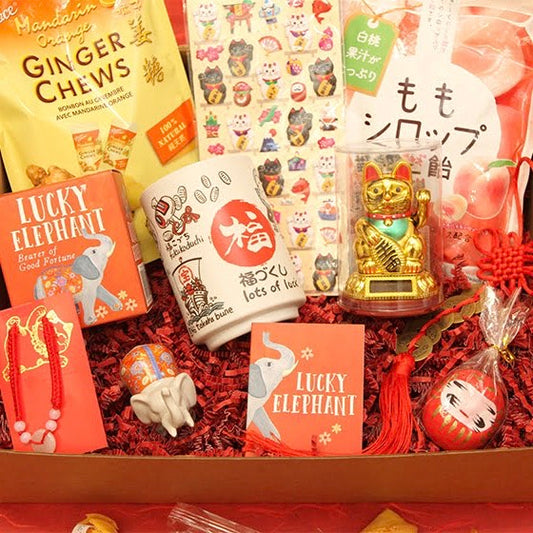 Box full of lucky items including orange ginger chews, fortune cat stickers, peach candies, lucky elephant kit, lots of luck teacup, lucky cat figurine, mini plant daruma