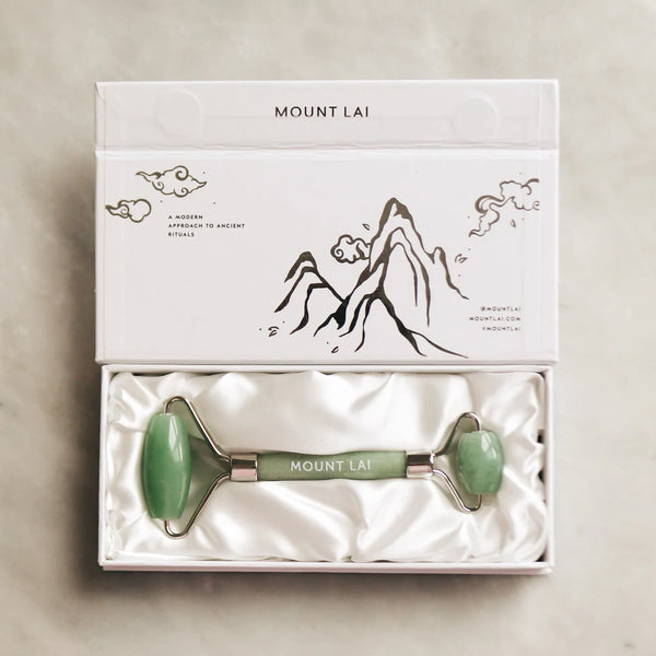 A Mount Lai jade facial roller presented in a white box with a drawing of mountains and the brand name. The roller has a green jade handle with a dual-sided design.