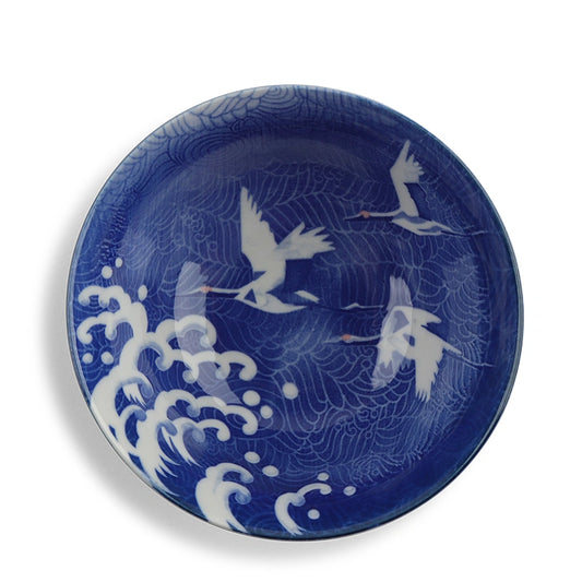 A round blue and white ceramic plate featuring a traditional Japanese-style design with white cranes flying over white waves on a deep cobalt blue background.