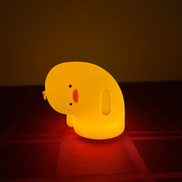 A cute, soft silicone duck night light glowing with a warm yellow light, resting on a dark red textured surface against a dark brown background.