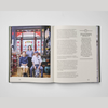 An open book spread photographed from above on a light gray background. The left page contains a color photograph of three people standing in a shop, and the right page contains multiple columns of black text on an off-white page.