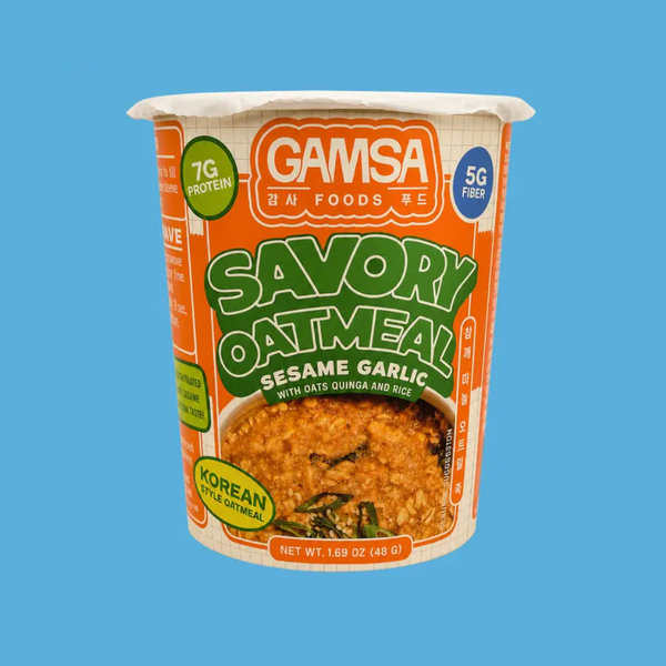 A cup of Gamsa Savory Oatmeal Sesame Garlic on a solid blue background. The packaging features orange and white colors with green text, and a photo of the prepared oatmeal with sesame garlic is shown on the front label.