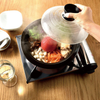 A hand lifts a clear lid off a black electric hot pot on a wooden table, with a steaming soup filled with vegetables and tofu inside.