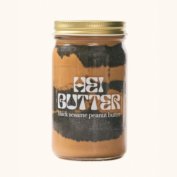 A glass jar of Hei Butter black sesame peanut butter with a gold metal lid and a black-and-white label, centered on a plain off-white background.