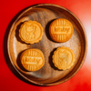 Four golden-brown mooncakes with embossed cat faces and the brand name 'kitsby' on two of them, arranged on a round wooden plate against a solid red background.