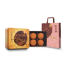 Four round Chinese mooncakes displayed in an open round tin next to a pink gift bag, with a matching closed tin behind them on a plain white background.
