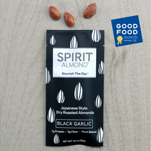 A single packet of Spirit Almond Black Garlic dry roasted almonds lies on a light wood surface, with three whole almonds placed above the package. A Good Food Awards Winner 26 badge appears in the upper right corner.