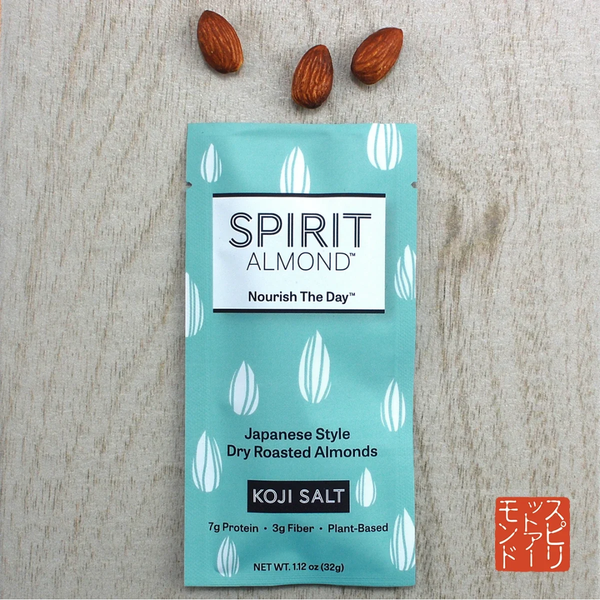 A package of Spirit Almond Koji Salt dry roasted almonds sits on a light wood background with three whole almonds placed above the package.